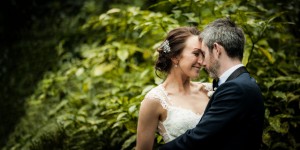 soft evening light illuminates the bride and groom cuddling in close and smiling at one another with green beautiful green bushes in the background