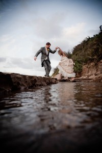 groom helps bride over stream at beach wedding